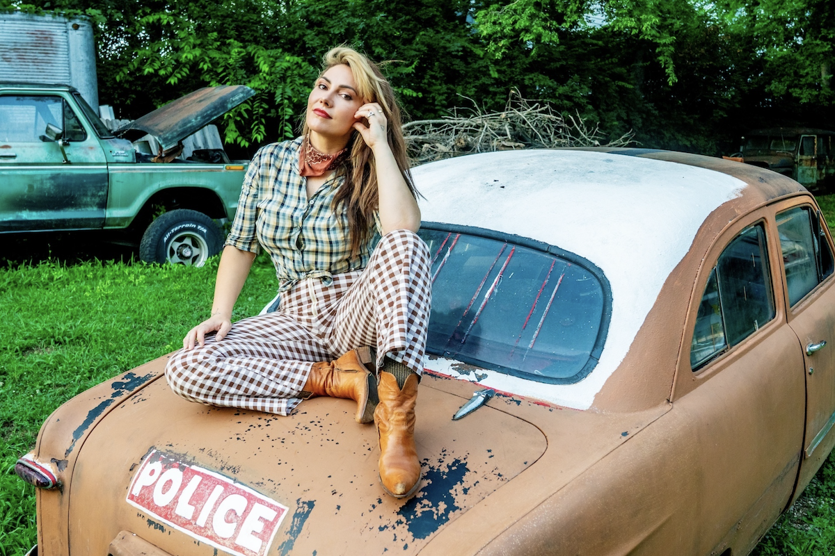 Australian singer Ella Hooper wearing cowboy boots and plaid pants, sitting on the trunk of a vintage car in an outdoor field.